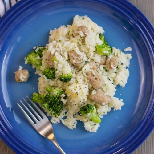 A blue plate holds a delightful One Pot Sausage Casserole with rice, broccoli, and sausage pieces. A fork is poised beside the dish, ready for a flavorful bite. In the backdrop, a striped cloth and wooden spatula add a rustic touch.