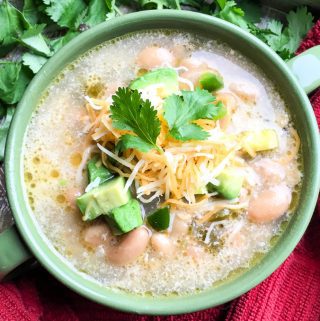 A bowl of slow cooker white chicken chili garnished with cheese, avocado, and cilantro on a red cloth.