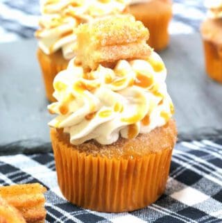 Churro Cupcakes Recipe 1 A decorated churro cupcake with a swirl of frosting and a cinnamon stick topping, displayed on a checkered cloth.