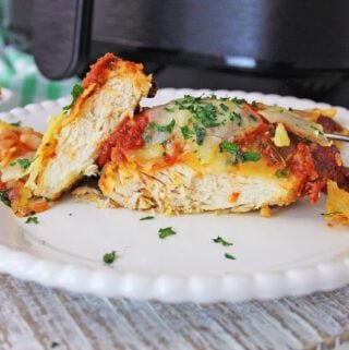 A plate with a sliced Air Fryer Chicken Parmesan topped with sauce and herbs, alongside melted cheese, with a fork and an air fryer in the background.