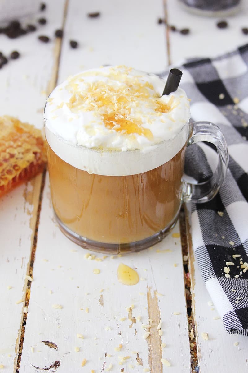 Glass mug of toasted coconut honey latte topped with whipped cream and caramel drizzle on a white wooden surface.