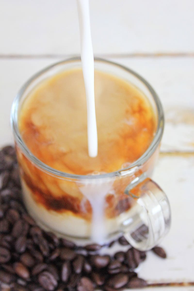 Pouring milk into a glass of Toasted Coconut Honey Latte surrounded by coffee beans.