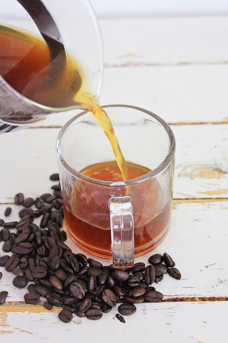 Pouring hot Toasted Coconut Honey Latte into a glass cup surrounded by coffee beans on a wooden surface.