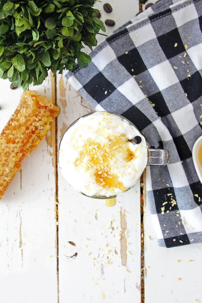 Top-down view of a cup of Toasted Coconut Honey Latte with whipped cream and zest on a white rustic wooden table, accompanied by honeycomb pieces and a checkered napkin.
