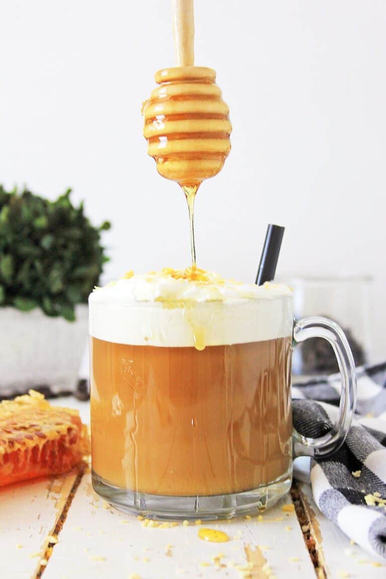 Honey drizzling onto a frothy cup of a Toasted Coconut Honey Latte with a backdrop of a light, bright kitchen setting.
