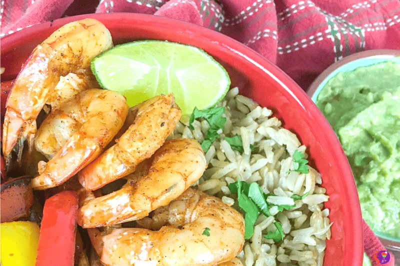 A bowl of shrimp fajita goodness with rice, sliced red and yellow peppers, topped with herbs. A lime wedge awaits on the side, while guacamole sits in a separate dish. A red checkered cloth completes the scene.