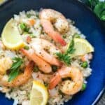 A blue bowl filled with prawn and pea risotto, garnished with lemon wedges and parsley, on a striped cloth background.
