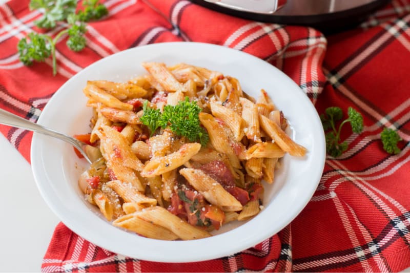 A bowl of Instant Pot penne pasta with tomato sauce and a garnish of parsley on a red and white checkered tablecloth.