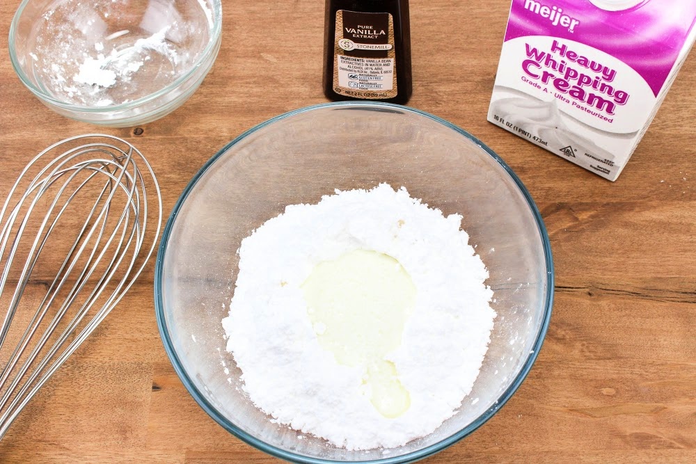 Ingredients for making whipped cream on a table include a bowl with powdered sugar and cream, vanilla extract, and a whisk. Perfect for topping your favorite desserts like caramel apple pull apart bread, all you need is some heavy whipping cream to whip up this delightful treat.