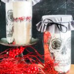Two jars, adorned with skull illustrations, stand on a table like mysterious Skull Cookie Mix in a Jar. One jar is perched on a pedestal, surrounded by red berries and twigs, wrapped in gray fabric for an eerie yet delightful touch.