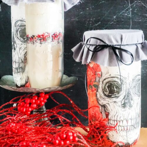 Two jars, adorned with skull illustrations, stand on a table like mysterious Skull Cookie Mix in a Jar. One jar is perched on a pedestal, surrounded by red berries and twigs, wrapped in gray fabric for an eerie yet delightful touch.
