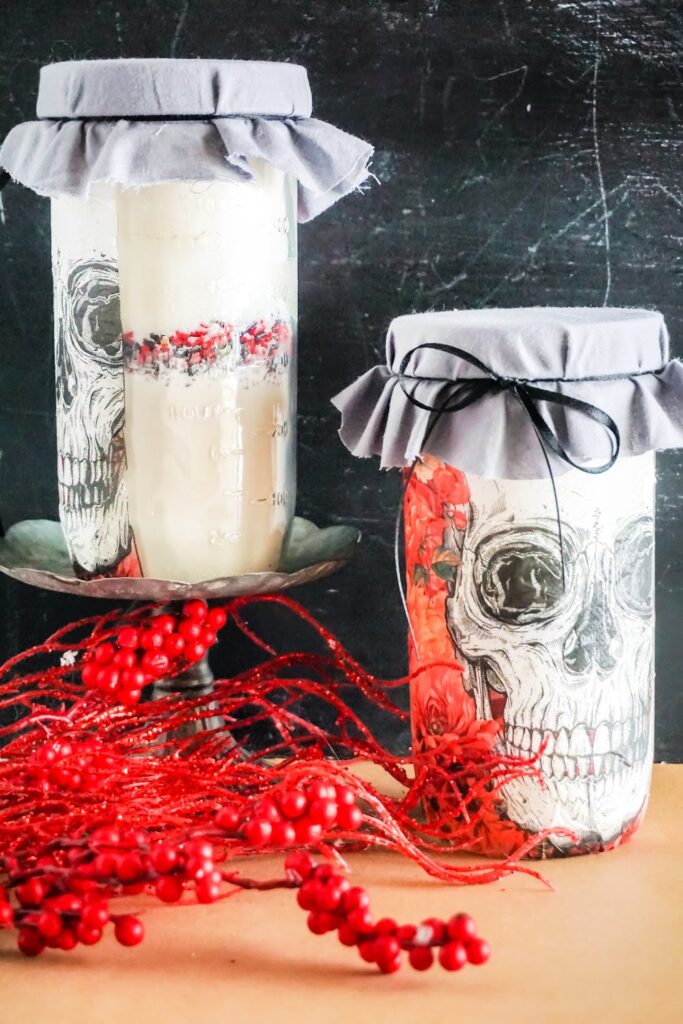 Two jars with skull designs and gray fabric lids sit elegantly. One jar, a Skull Cookie Mix in a Jar, boasts layers of ingredients. Red berries and twigs artfully scatter before them against a dark background, adding an intriguing touch to the mysterious scene.