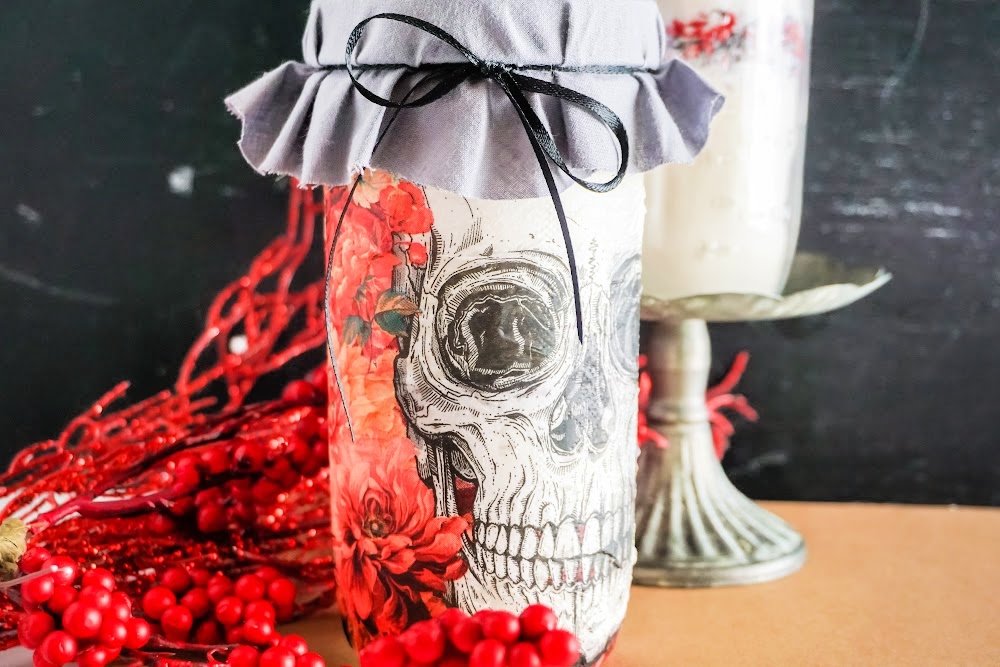 Decorative jar with a skull cookie mix design, gray fabric cover tied with a black ribbon. Red flowers and berries are visible around the base, adding a touch of warmth. A candle holder glows softly in the background.