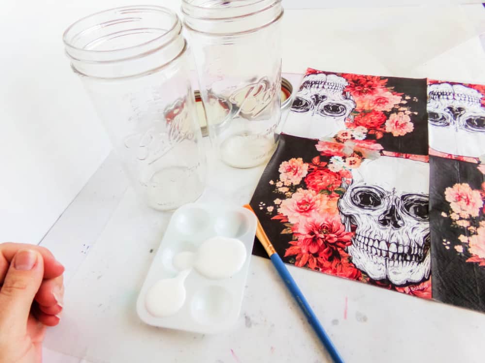 Decorative jar featuring a skull illustration, reminiscent of a Skull Cookie Mix in a Jar, topped with gray fabric and bow. It's surrounded by vibrant red flowers and berries, all set against a dark background for an edgy yet elegant look.