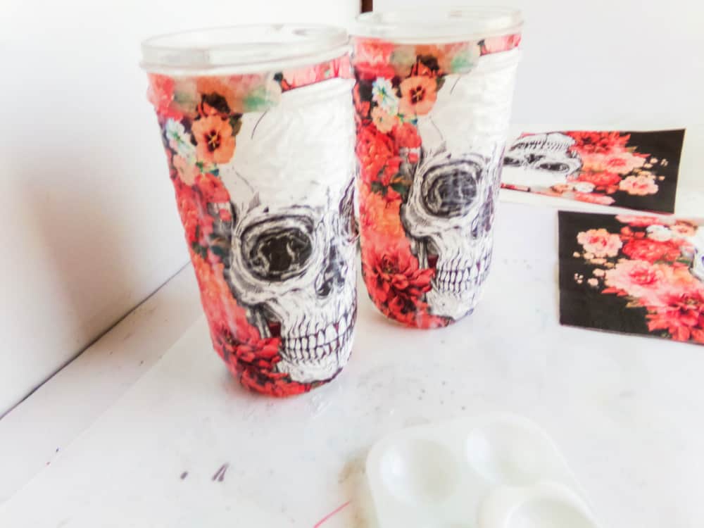 A hand pours black, red, and orange sprinkles into a glass jar labeled Skull Cookie Mix in a Jar. Nearby, another jar holds powder while a bowl of flour sits with a measuring spoon, ready to create spooky treats.