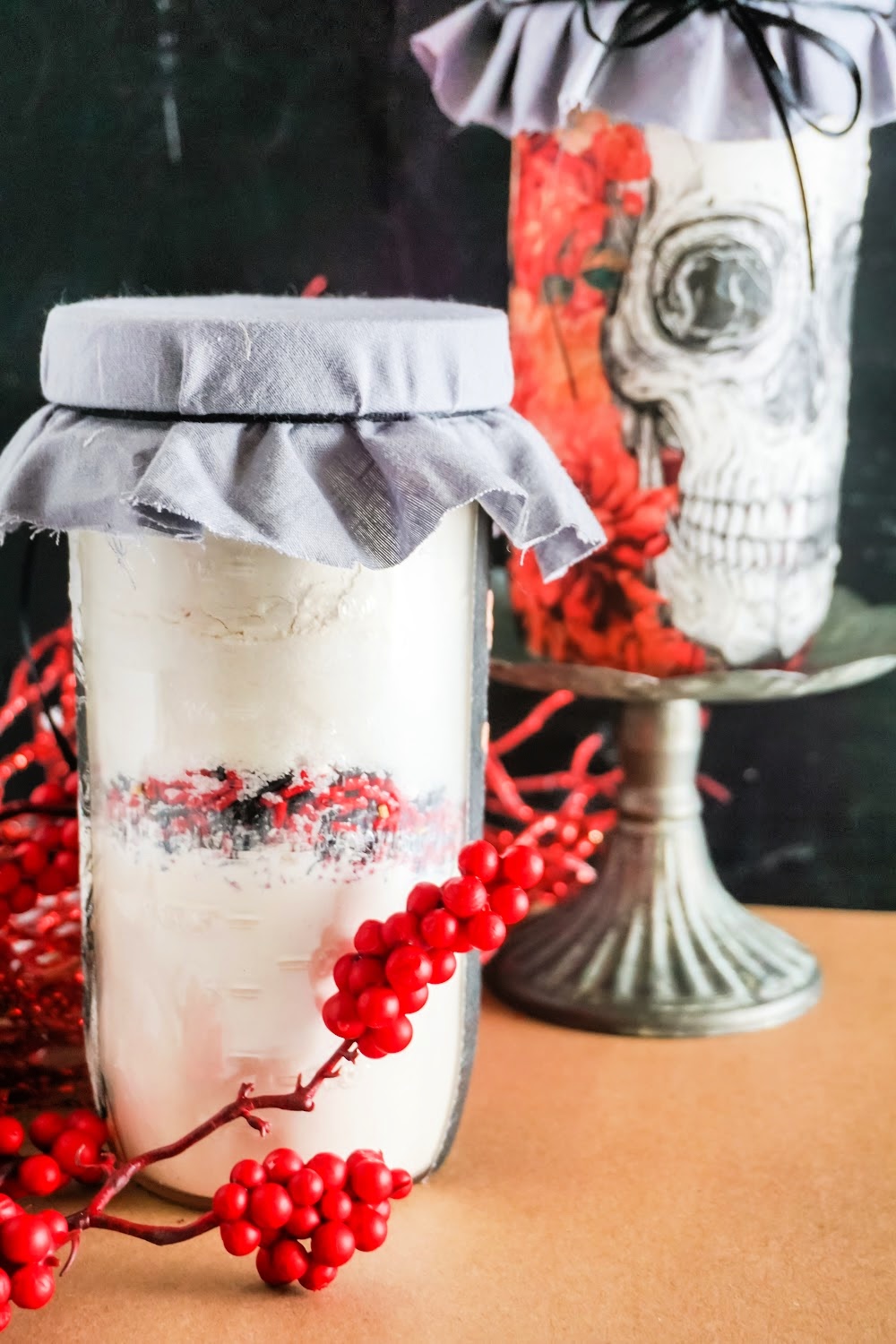 Two glass jars with fabric covers create a striking scene. One is filled with a white substance and red berries; the other showcases a clever skull cookie mix in a jar design. Red berries are artfully scattered, all set against a dark, moody background.