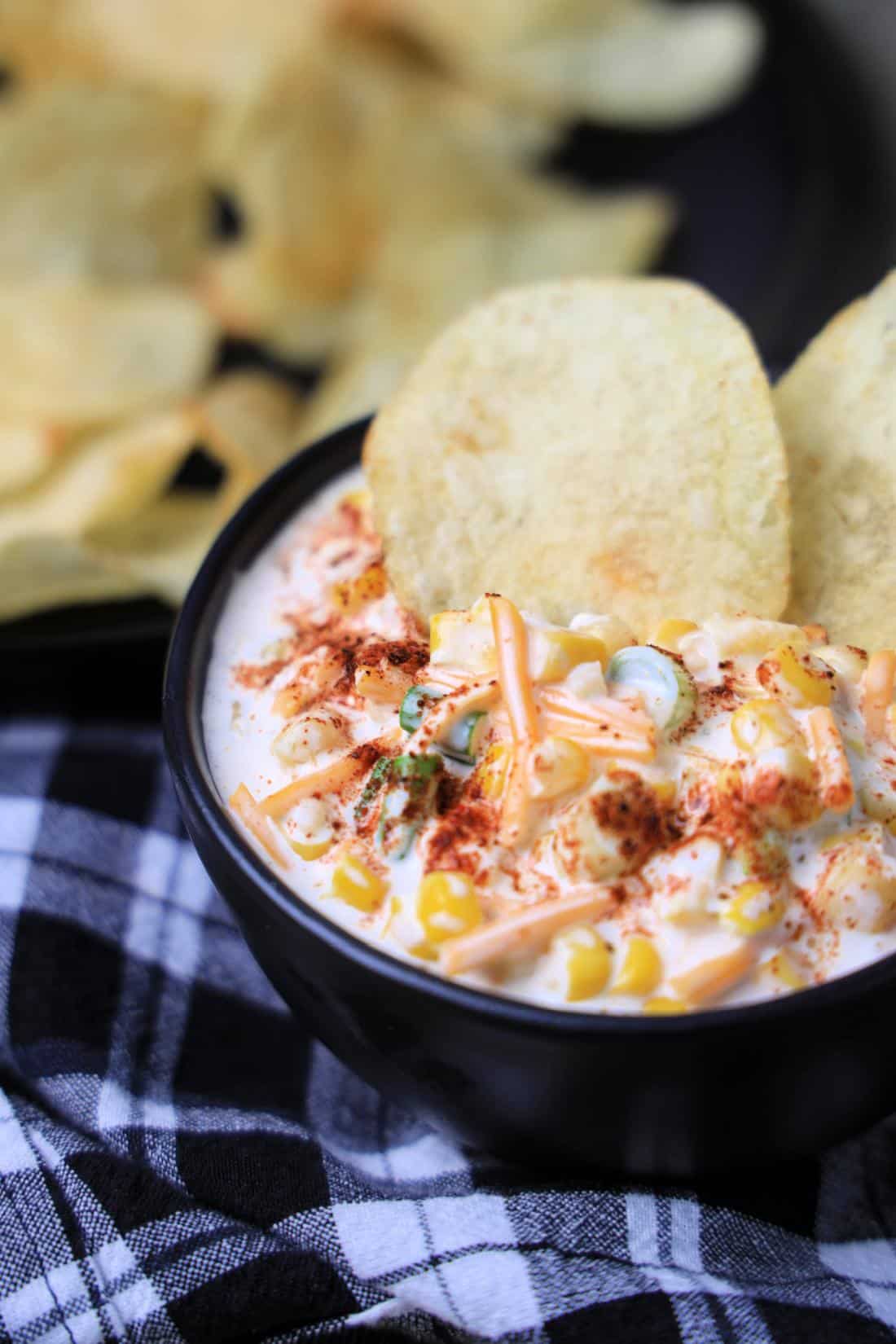 A bowl of Southwestern Corn Dip garnished with shredded cheese and spices, served with potato chips on the side.