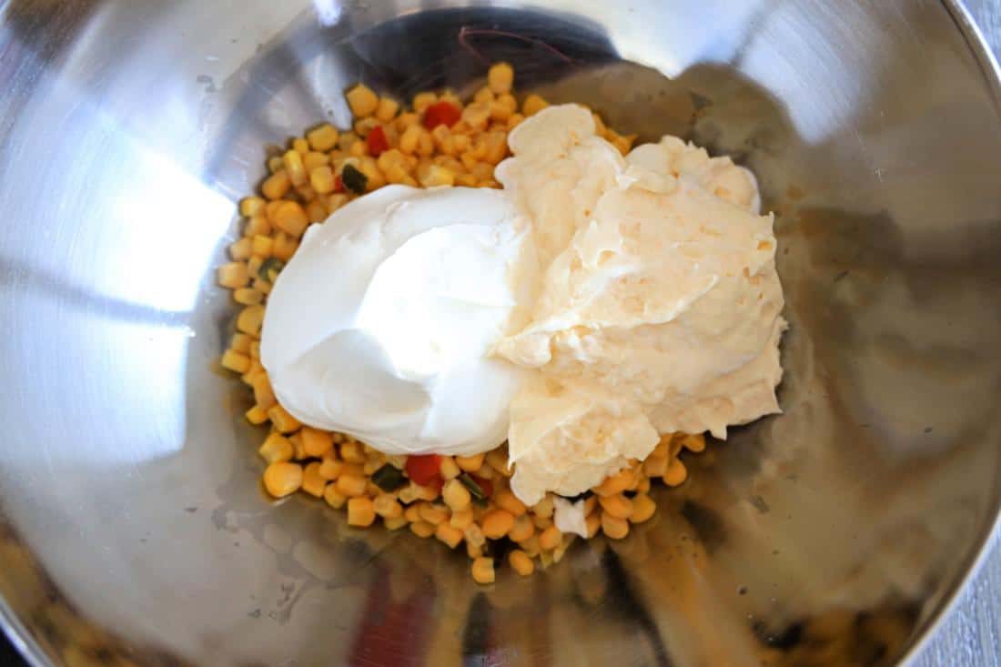Ingredients for Southwestern Corn Dip in a mixing bowl, including corn, red peppers, and two types of cream or soft cheese.