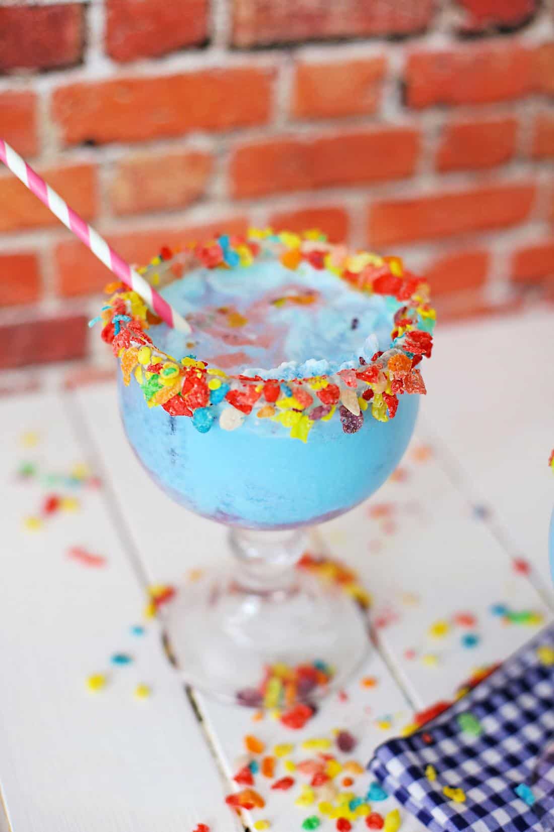 A colorful Fruity Pebbles Margarita with a candy-rimmed glass and a striped straw placed on a white table with scattered candies against a brick wall background.