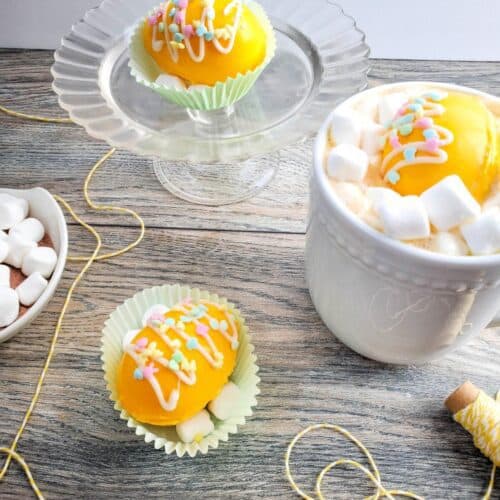 The Best Yellow Easter Egg Hot Cocoa Bombs 16 A bright and festive dessert setup featuring two yellow frosted cupcakes with colorful sprinkles, one on a glass stand and another in a paper cup, accompanied by a mug of hot beverage topped with Easter Egg