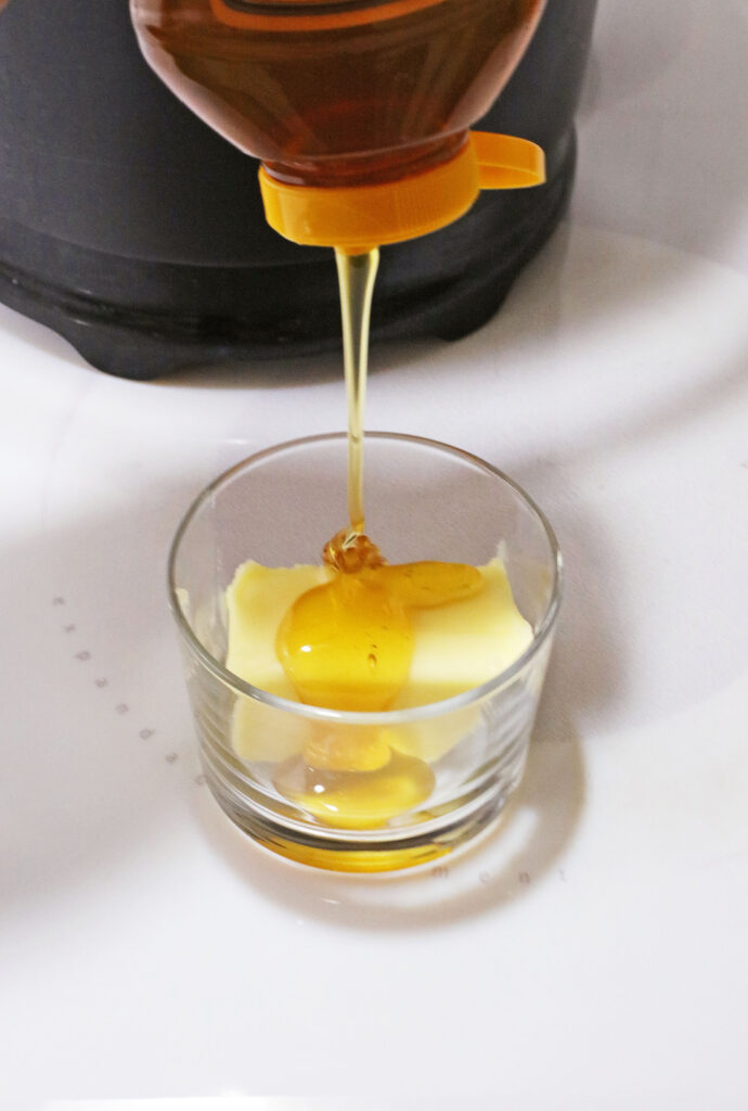 Copy Cat Chick-Fil-A Chicken Mini's 10 Golden honey cascades from a bottle into a glass bowl, enveloping a piece of butter on a pristine white surface—a scene reminiscent of the warm, fluffy delight found in Chick-Fil-A Chicken Minis.