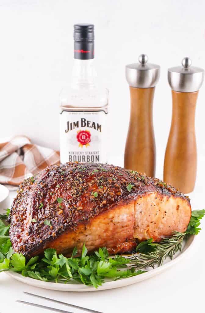 Roasted herb-crusted glazed ham served on a plate with parsley, accompanied by a bottle of Jim Beam bourbon, salt and pepper grinders in the background.