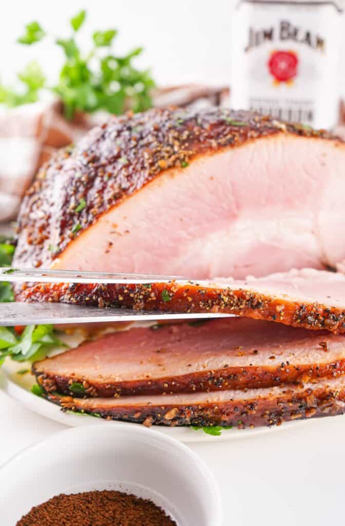 Slicing a pepper-crusted, bourbon glazed ham with visible seasoning next to a bottle of bourbon.