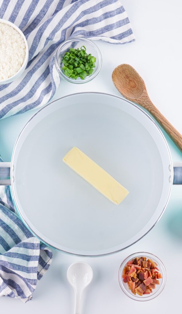 Butter in a frying pan surrounded by ingredients like green onions, crumbled bacon, and flour, perfect for starting a delicious Loaded Baked Mashed Potato Casserole. A striped cloth and wooden spoon lie nearby.