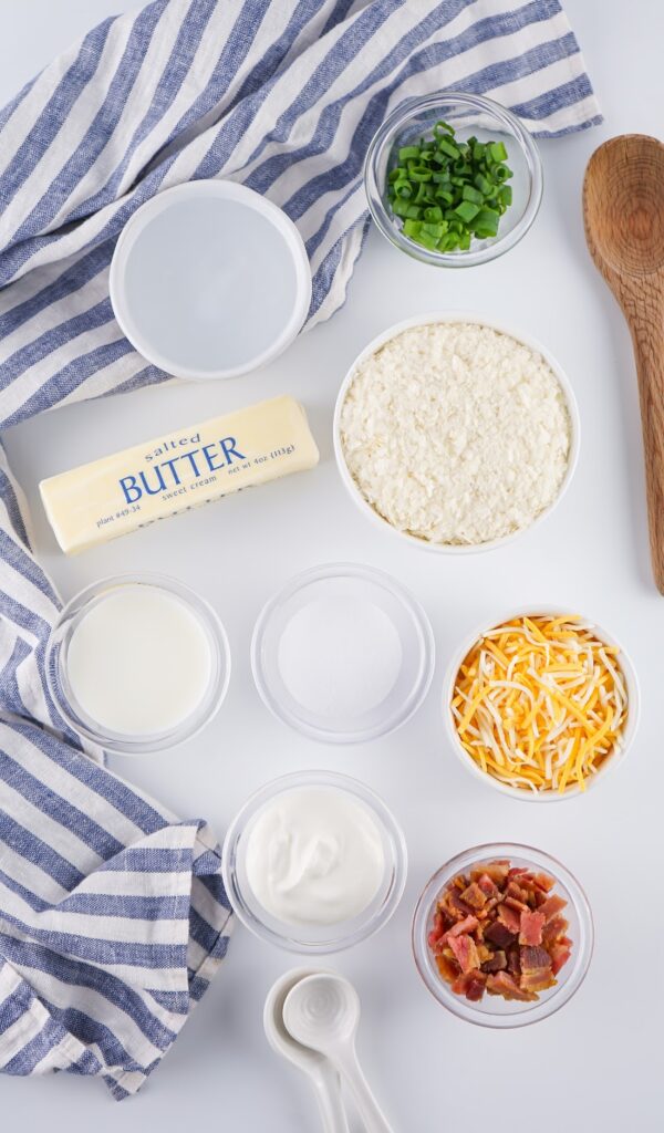 Overhead view of ingredients for Loaded Mashed Potatoes: butter, flour, milk, water, sugar, sour cream, chopped bacon, shredded cheese, and green onions. All carefully arranged on a striped cloth with a wooden spoon and measuring spoon nearby.