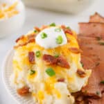 A plate showcasing sliced ham alongside loaded mashed potatoes, topped with cheese, bacon bits, sour cream, and chopped green onions. A dish of shredded cheese is in the background. This hearty meal highlights the irresistible charm of Loaded Baked Mashed Potato delight.