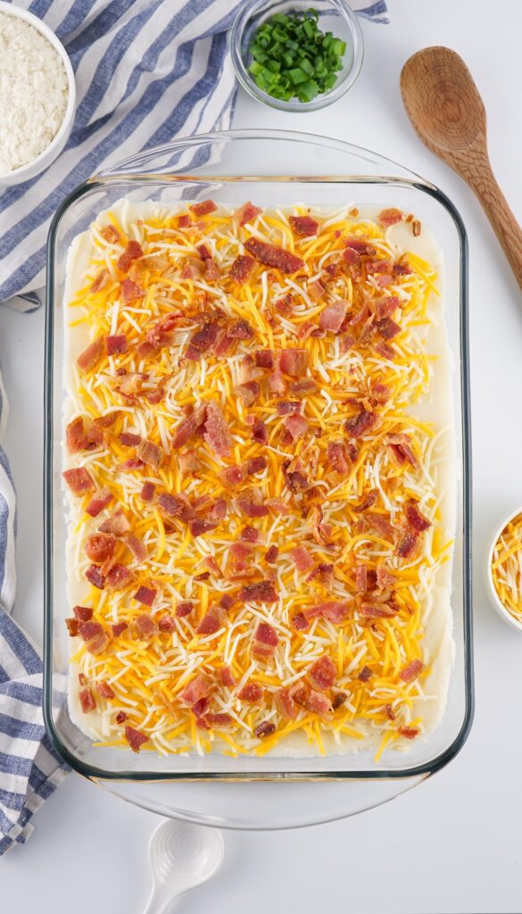A glass dish showcases a layered casserole, topped with cheese and crispy bacon pieces. Nearby, find a striped cloth, a bowl of scallions, wooden spoon, and cheese in a small bowl—perfect accompaniments to creamy loaded baked mashed potatoes.