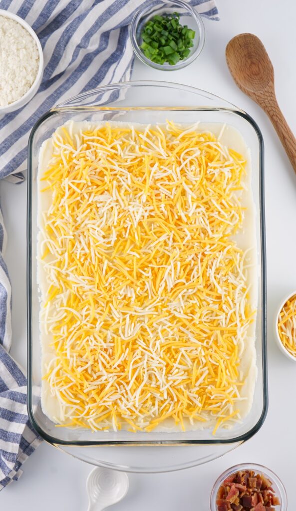 A glass baking dish brimming with creamy mashed potatoes topped with shredded cheese, surrounded by chopped green onions and bacon bits, rests beside a wooden spoon. A striped cloth adds a cozy touch.