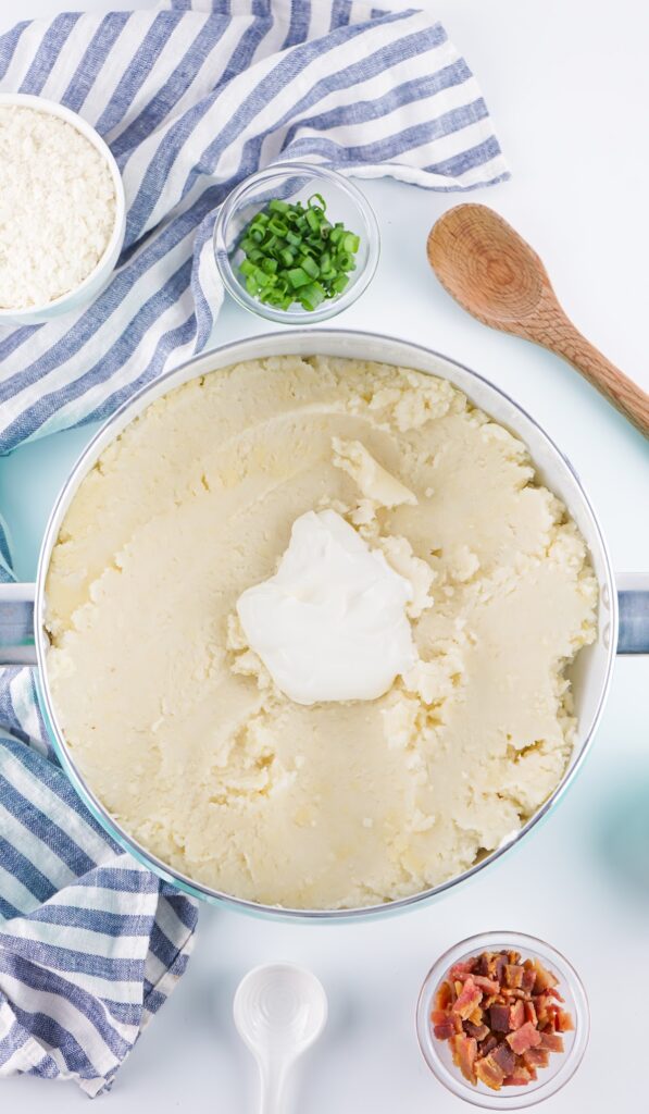 A pot of creamy mashed potatoes topped with sour cream, surrounded by green onions, bacon bits, a wooden spoon, and flour on a striped cloth.
