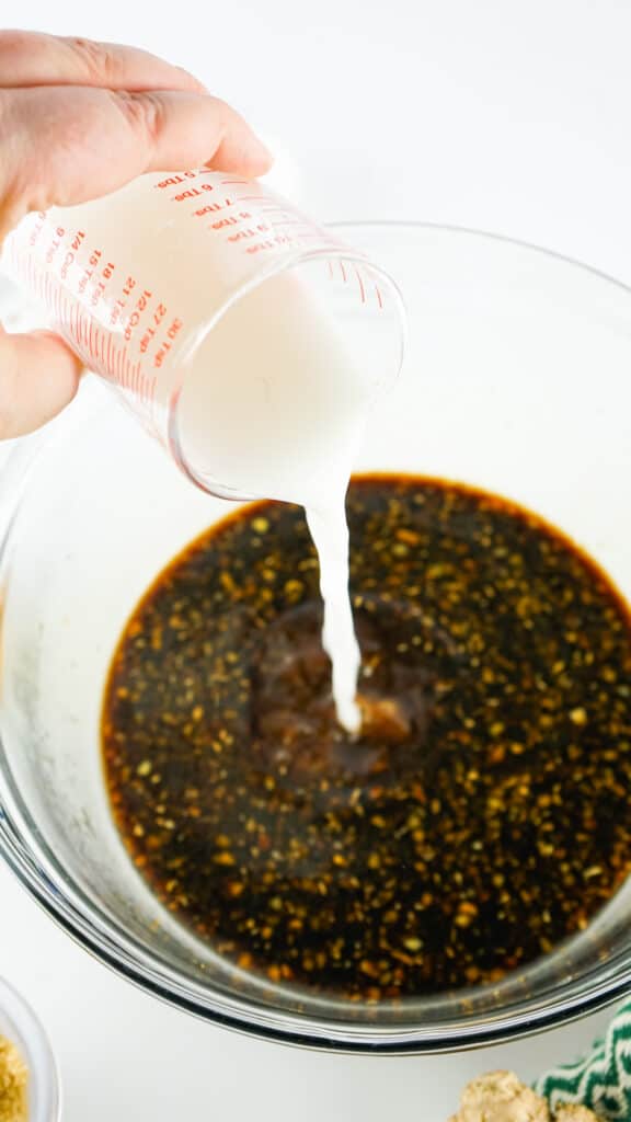Pouring milk from a measuring cup into a bowl of Teriyaki sauce with herbs and spices on a white background.