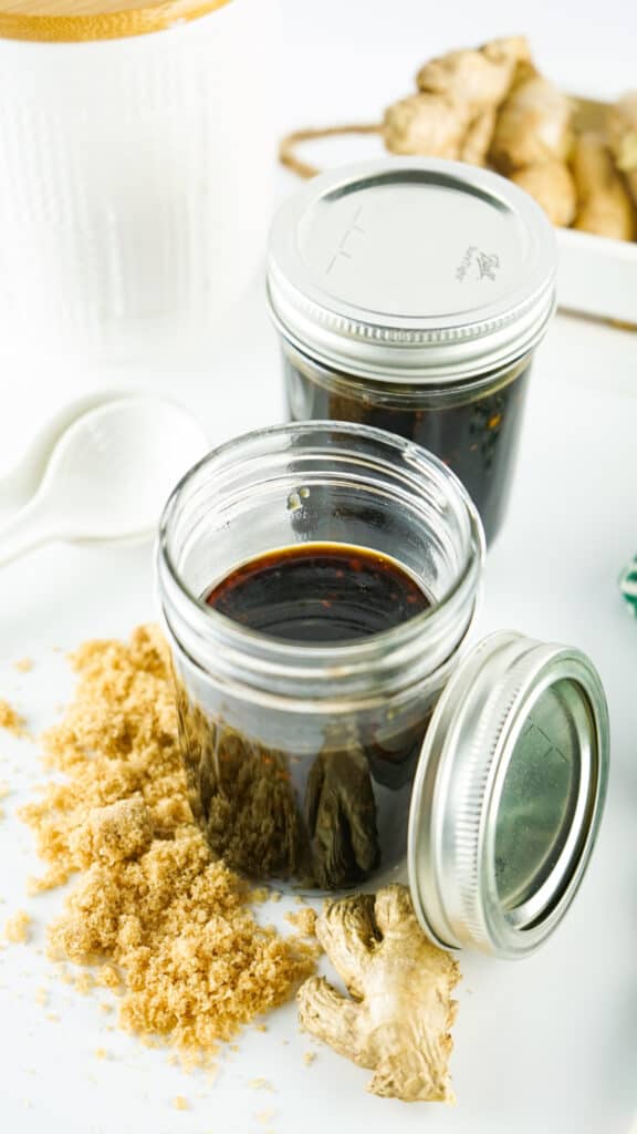 Two jars of homemade teriyaki sauce on a white surface, surrounded by fresh ginger roots and brown sugar.
