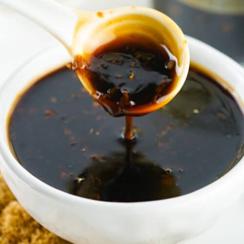 Spoon pouring thick teriyaki sauce into a white bowl, with granulated sugar and ginger root visible in the background.