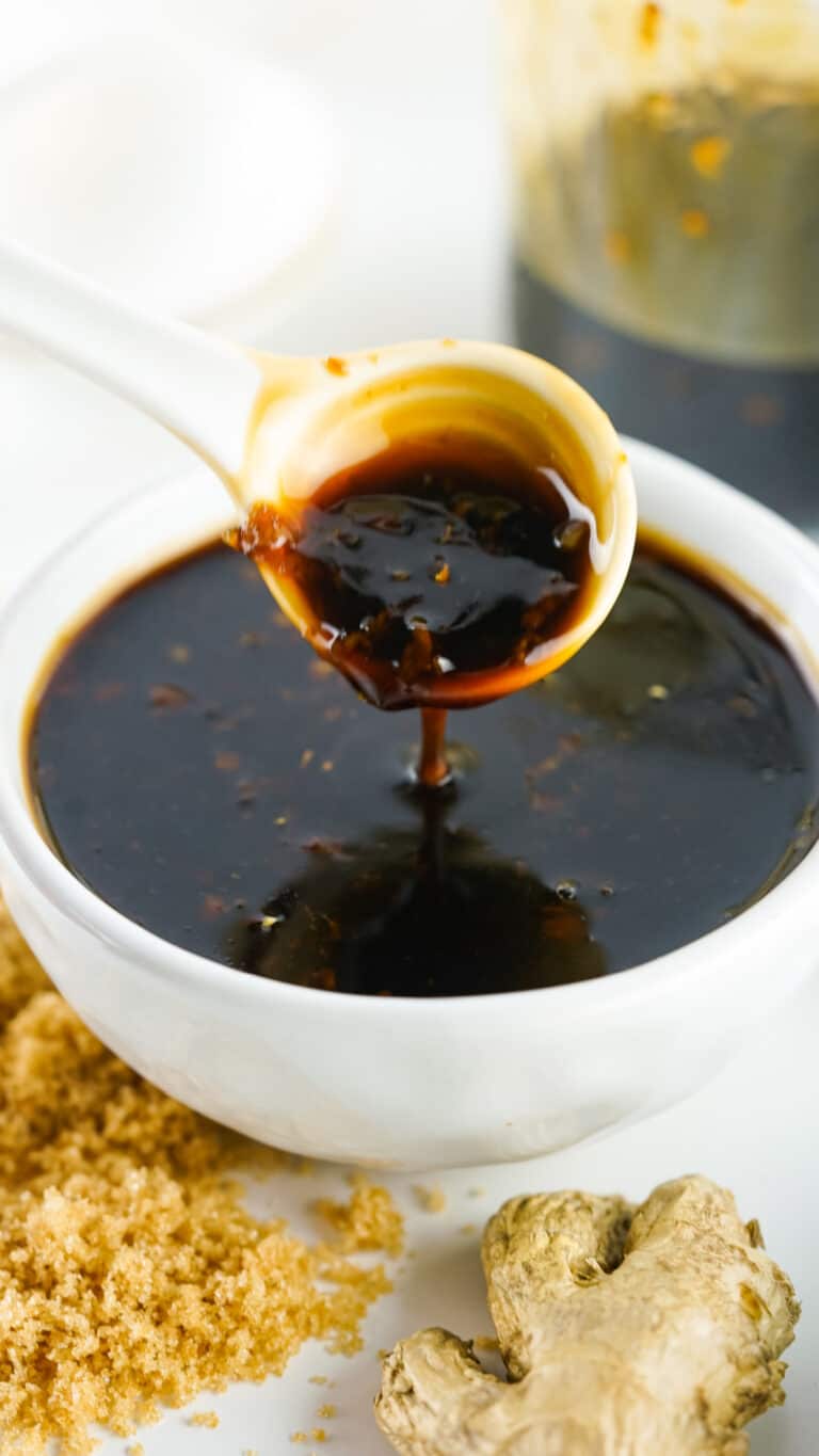 Spoon pouring thick teriyaki sauce into a white bowl, with granulated sugar and ginger root visible in the background.