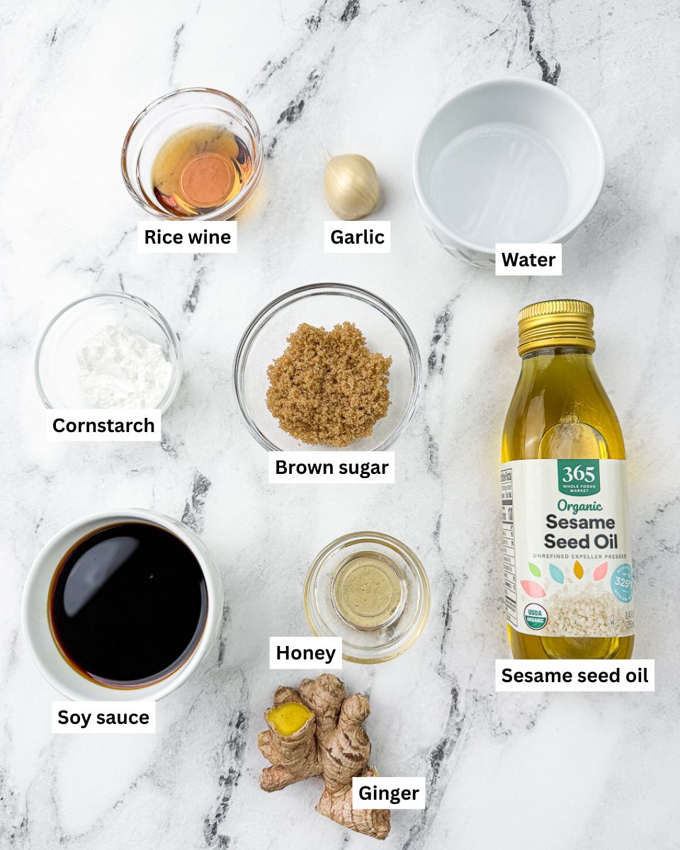 Ingredients for teriyaki sauce layed on countertop in various bowls: rice wine, garlic clove, water, cornstarch, brown sugar, sesame seed oil, soy sauce, honey, ginger