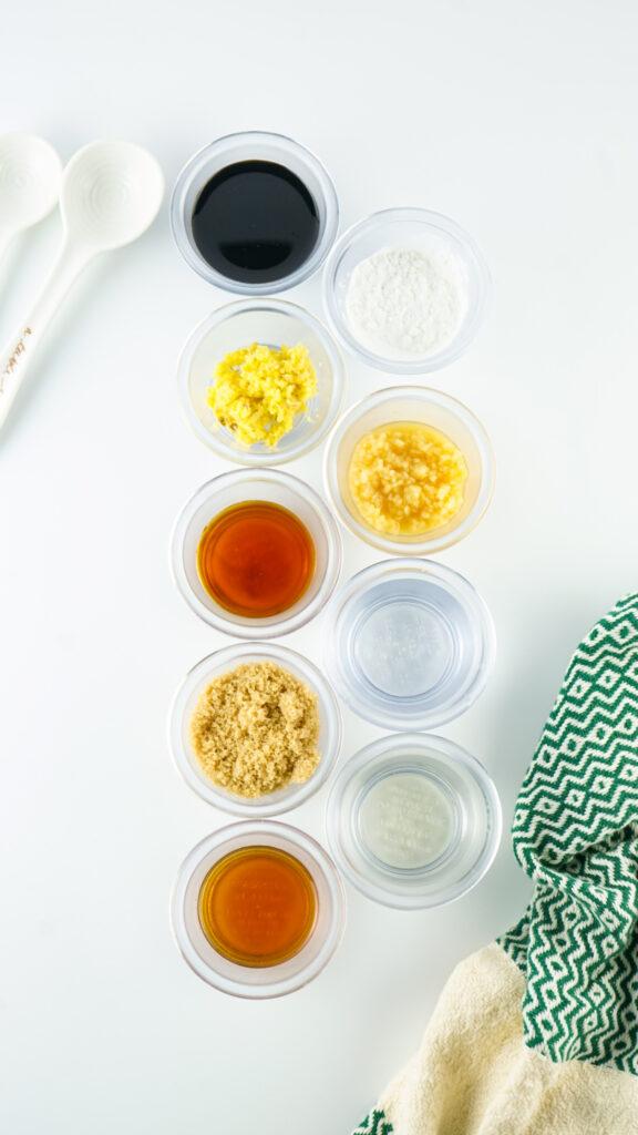 Various cooking ingredients in small clear containers, including spices and liquids for homemade Teriyaki sauce, arranged neatly on a surface beside white measuring spoons and a green patterned towel.