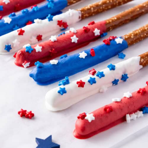 Firecracker Pretzel Rods coated with red, white, and blue chocolate and decorated with star-shaped sprinkles are arranged on a white surface, making them the perfect easy party snacks.