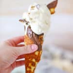Indulge in a delightful ice cream cone featuring a scoop of no-churn vanilla ice cream, adorned with chocolate pieces and a luscious chocolate drizzle, all elegantly set against a checkered cloth on a wooden background. Perfectly reminiscent of Ben & Jerry’s Copycat No Churn Americone Dream Ice Cream.
