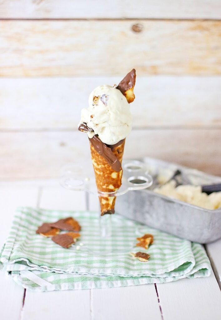 A waffle cone brimming with chocolate and vanilla no-churn ice cream rests on a stand. A green striped cloth lies beneath, while a metal tray with more Ben & Jerry's creations lurks in the background, tempting every sweet tooth.