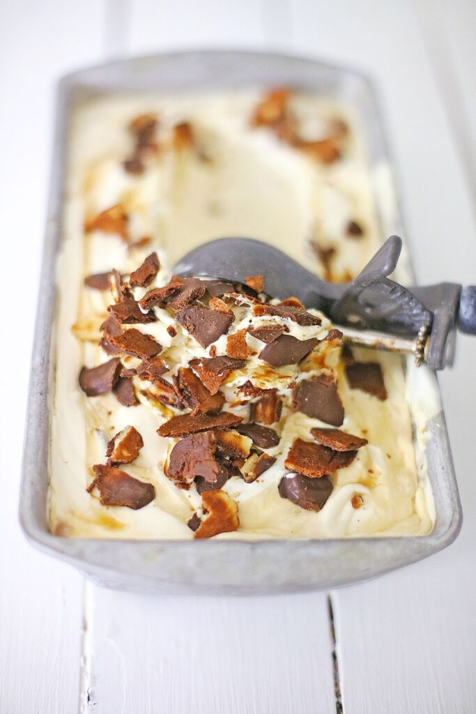 A pan of Ben & Jerry's copycat No Churn Americone Dream Ice Cream topped with chocolate and caramel pieces, with an ice cream scoop resting on top.