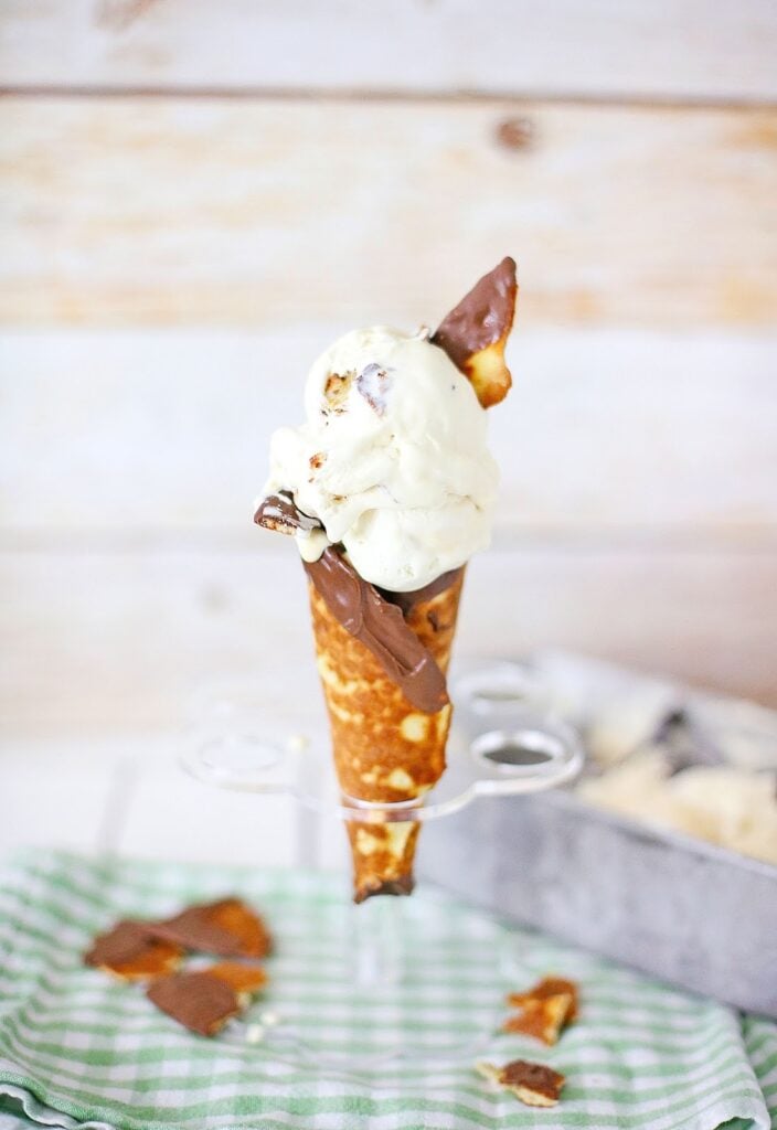 An ice cream cone featuring chocolate and caramel sits on a stand, surrounded by broken cone pieces on a green checkered cloth. Experience the taste of Ben & Jerry's Copycat No Churn Homemade Americone Dream Ice Cream in this delightful scene.