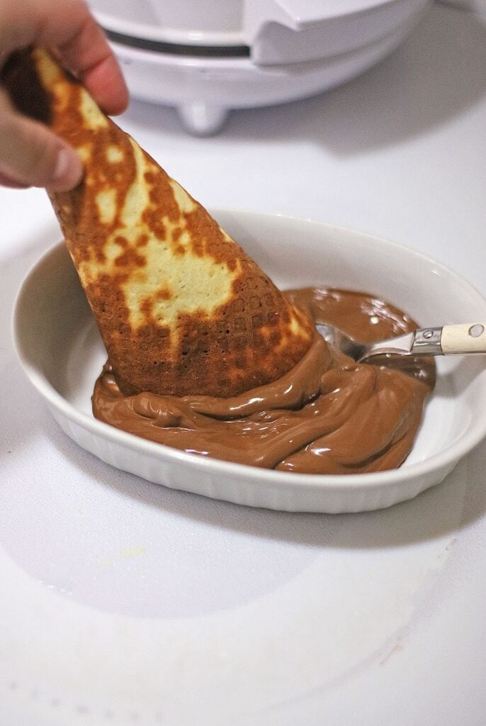 A hand dips a folded crepe into a dish of thick chocolate sauce with a spoon on the side, reminiscent of indulgent flavors found in Ben & Jerry's Copycat No Churn Homemade Americone Dream Ice Cream.