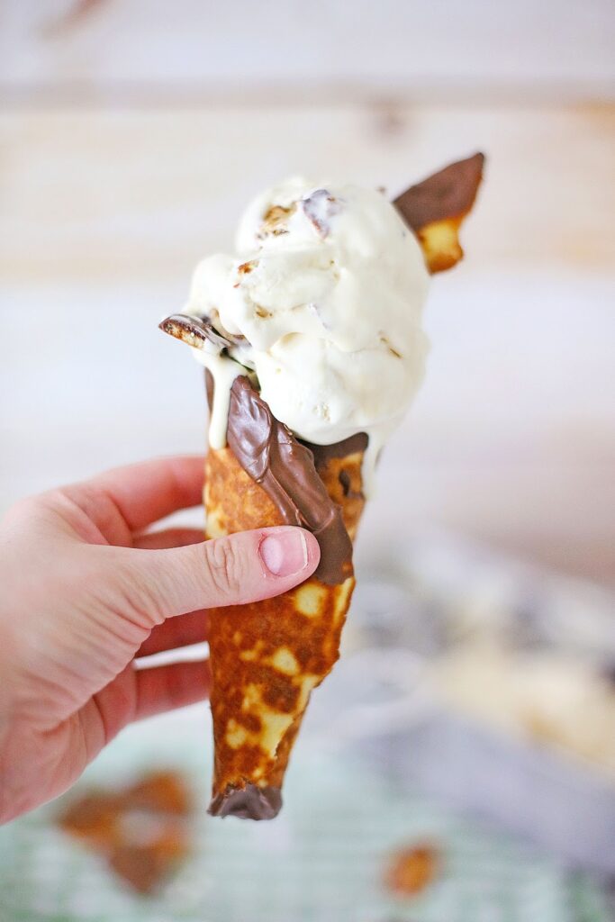 A hand holds a chocolate-dipped waffle cone filled with creamy vanilla ice cream reminiscent of Ben & Jerry's Copycat No Churn Homemade Americone Dream Ice Cream, elegantly topped with a chocolate piece.