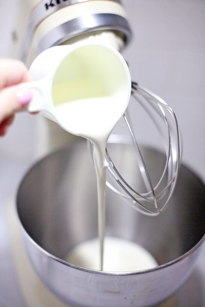 A person pouring cream into a stand mixer bowl with a whisk attachment, preparing for a batch of Ben & Jerry's Copycat No Churn Homemade Americone Dream Ice Cream.