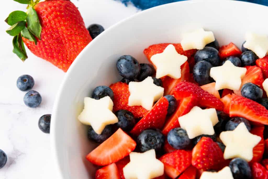 A white bowl filled with cut strawberries, blueberries, and star-shaped pieces of fruit creates a delightful Star Spangled Fruit Salad; additional strawberries and blueberries are placed outside the bowl on a marble surface.