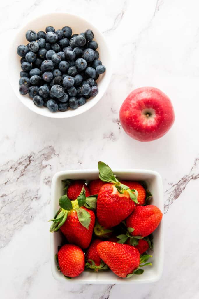 A white bowl of blueberries, a red apple, and a white square dish of strawberries sit on a white marble surface, coming together to create the perfect Red White and Blue Fruit Salad.