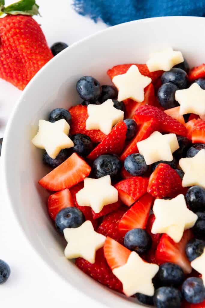 A white bowl contains a Star Spangled Fruit Salad with sliced strawberries, blueberries, and star-shaped pieces of melon. A whole strawberry and a few scattered blueberries are visible near the bowl.